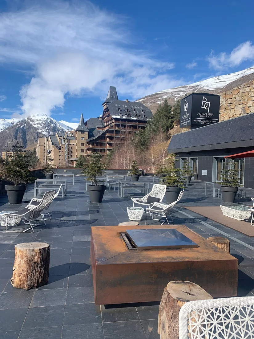 The rooftop terrace of the AC Baqueira Autograph Collection with a steel fire pit and loungers, the hotel and snowy Pyrenees behind, and 'AC Baqueira Ski Resort — Autograph Collection' signage
