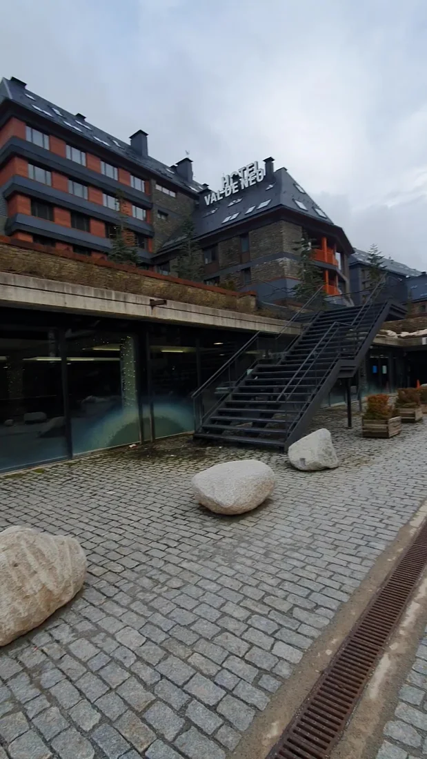 The stone-and-slate facade of Hotel Val de Neu with its rooftop 'HOTEL VAL DE NEU' sign, above a cobbled plaza at the Baqueira base