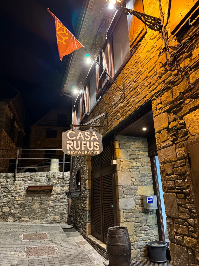 The lit wooden 'CASA RUFUS Restaurante' sign and Aranese flag on a stone building in Gessa at night
