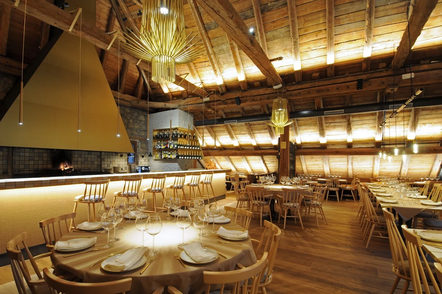 The wood-beamed dining room of La Borda Lobató, a converted stone barn with an open fire, wine bar and round tables set under pendant lights