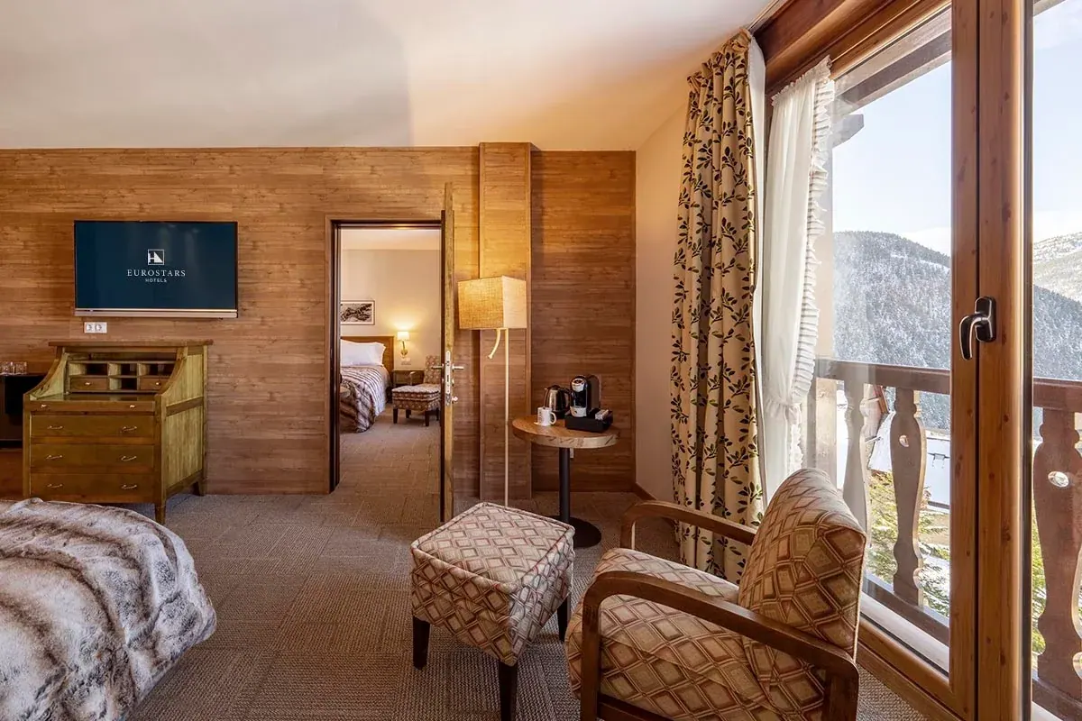 A wood-panelled Eurostars La Pleta suite with a balcony over the snowy mountains and the Eurostars logo on the room TV