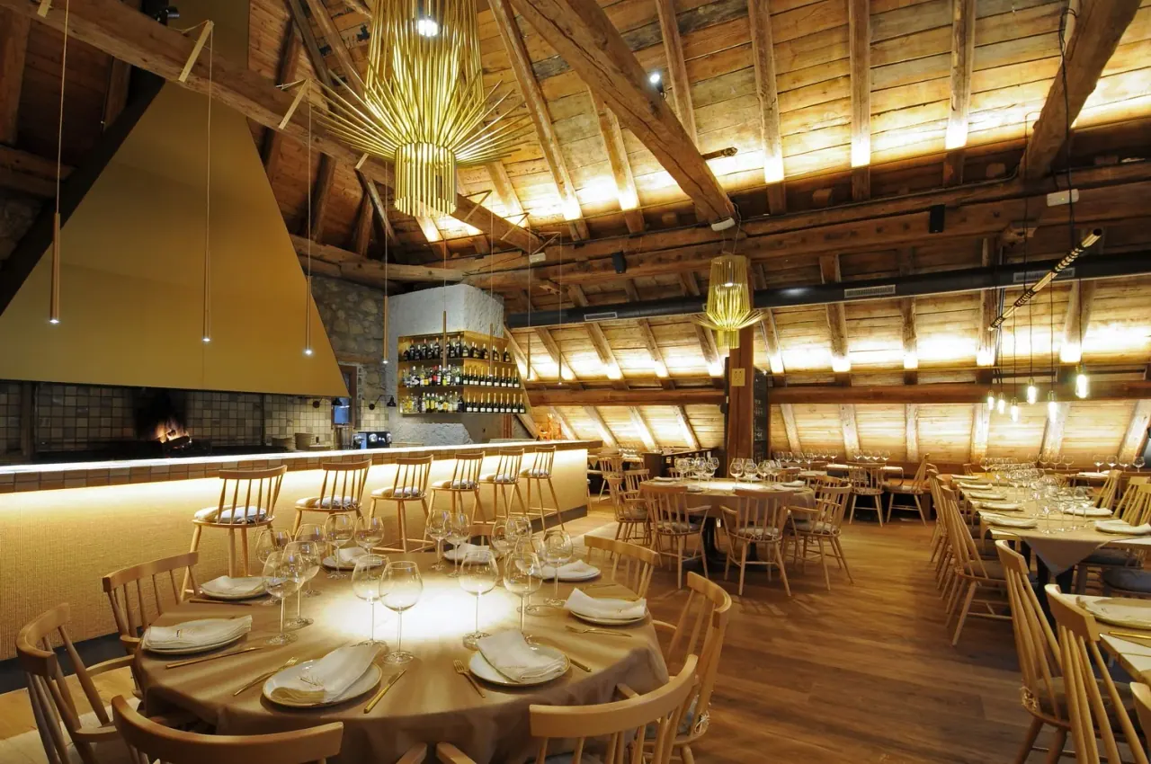 The wood-beamed dining room of La Borda Lobató, a converted stone barn with an open fire, wine bar and round tables set under pendant lights